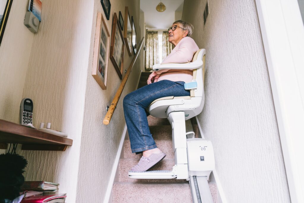 Elderly woman seated on a stairlift riding up a narrow residential staircase inside her home